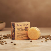 Tanamera Toning Green Coffee Body Soap shown with its kraft paper box, placed on a wooden surface with scattered coffee beans. The round soap bar is a warm golden-beige color, and the box is brown with black text and coffee bean imagery.